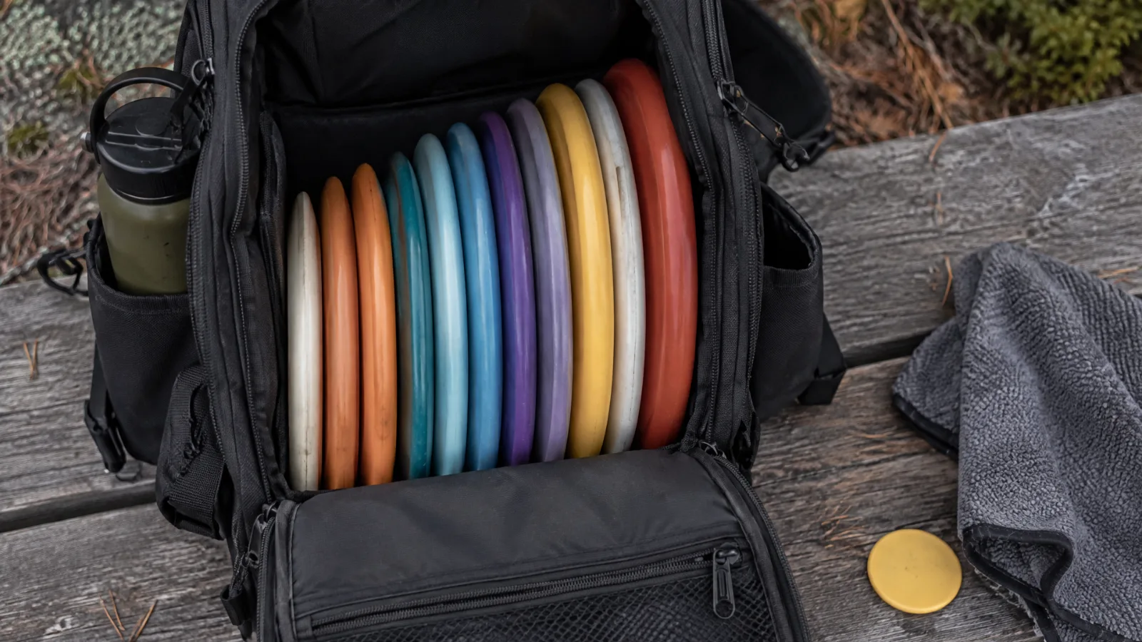 Åpen discgolf-bag på en benk i skogen med tolv disker i ulike farger, fra puttere til drivere, med vannflaske, håndkle og markør ved siden av