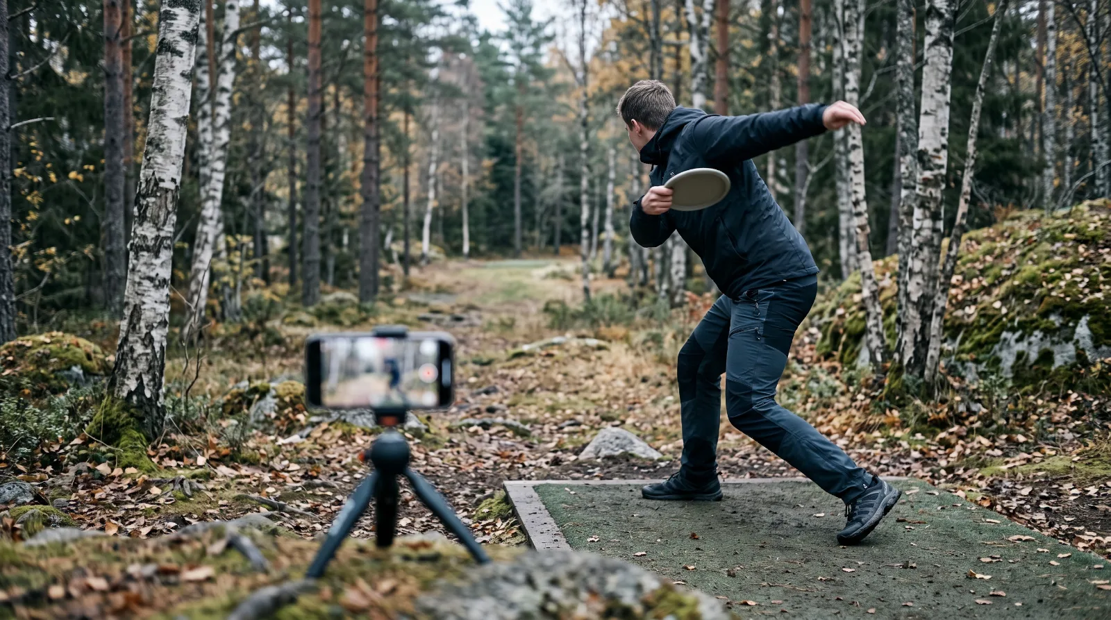 Spiller kaster backhand fra tee i skogen mens en mobil på stativ filmer kastet fra siden