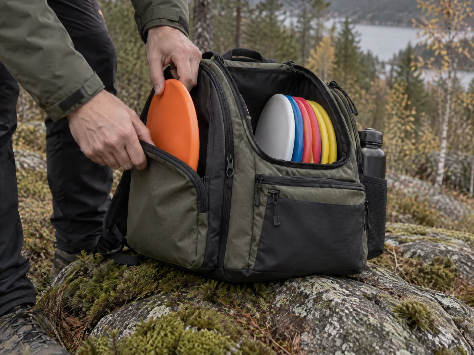 Spiller legger en oransje disk i en åpen discgolf-bag på en mosegrodd stein, med flere disker i bagen og et vann i bakgrunnen
